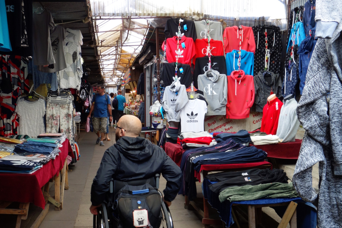 Bild vom Polenmarkt. Ein schmaler Gang mit Kleidungsständen recht und links. Ein Rollstuhlfaher fährt den Gang entlang.
