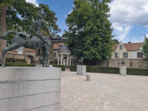 Statue eines nackten Reiters auf einem Pferd mit durchbrochener Oberfläche auf Betonpodest, im Hintergrund weitere Reiterstatuen auf Podesten.
