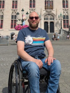 Mann im Rollstuhl lächelt in die Kamera vor historischen Gebäuden.
