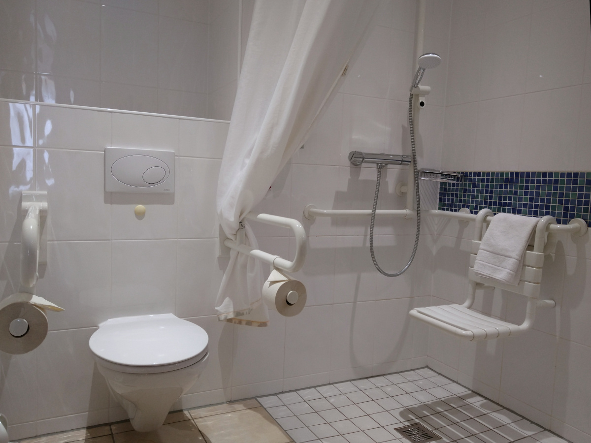 Behinderten-Toilette. Links Toilette mit 2 Stütz-Griffen. Rechts die stufenlos zugängliche Dusche mit Sitz an der Wand.