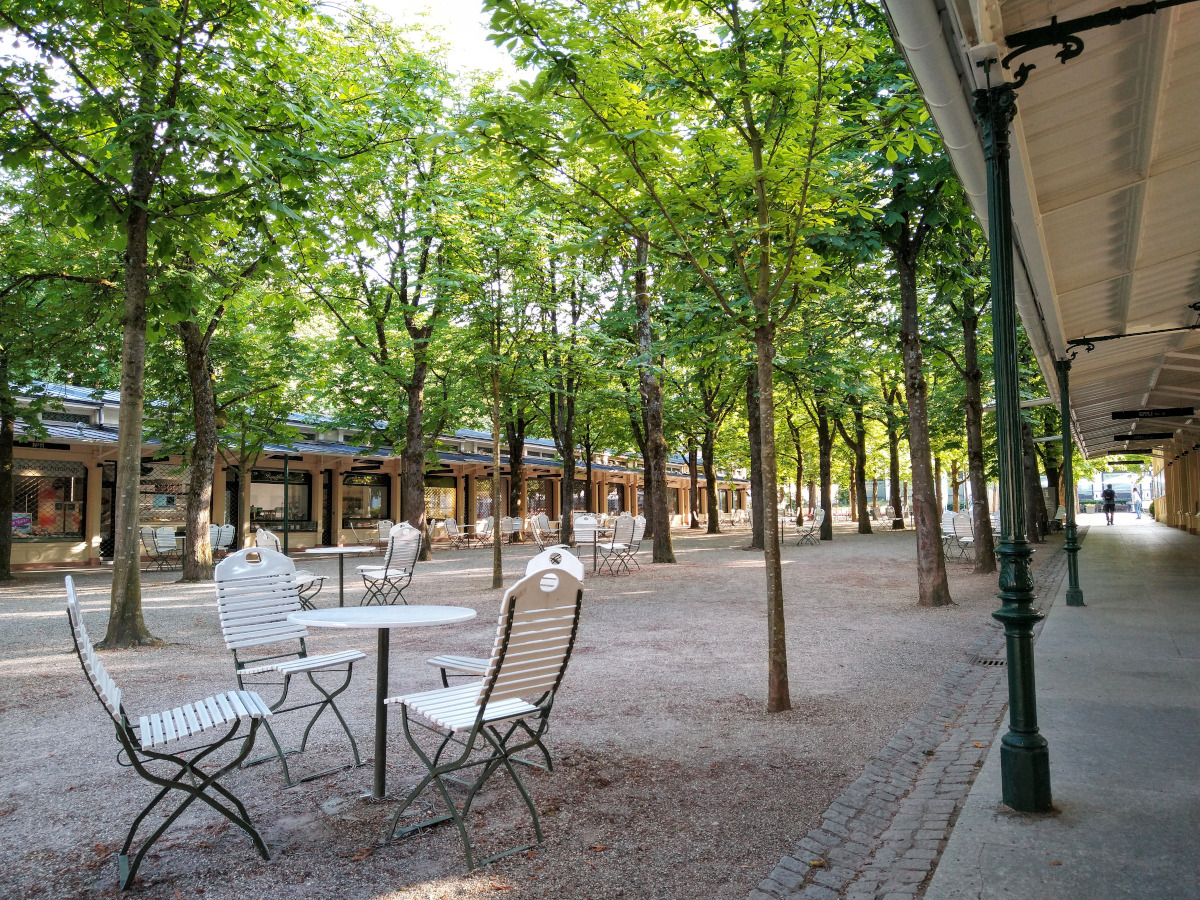 Platz mit Kies-Boden. Darauf eine 3-reihige Allee mit Bäumen. Im Schatten der Bäume Tische mit Stühlen. An den Seiten kleine Geschäfte.