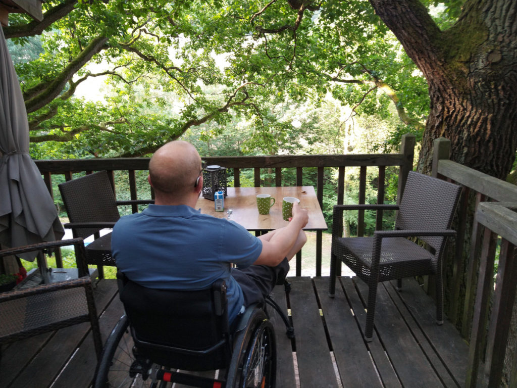 Rollstuhlfahrer auf einer Holz-Terrasse mit einem Tisch und Stühlen. Auf dem Tisch eine Tasse Kaffee. Im Hintergrund Blätter und Äste.