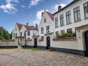 Mehrere kleine weiße Häuschen hinter brusthohen Mauern mit Eingangstüren, davor ein geteilter Weg mit ebenem Pflaster links und unebenem historischem Kopfsteinpflaster rechts.