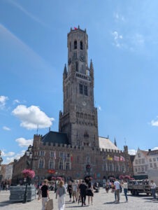 Eckiges dreistöckiges gemauertes Gebäude mit hohem, sich in drei Abschnitten verjüngender Turm in der Mitte, davor gepflasterter Platz mit vielen Menschen.