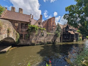 Kleiner Fluss, links von historischen gemauerten Häusern gesäumt, rechts ein über das Wasser ragender Baum. Links im Bild eine teilweise sichtbare gemauerte Brücke.