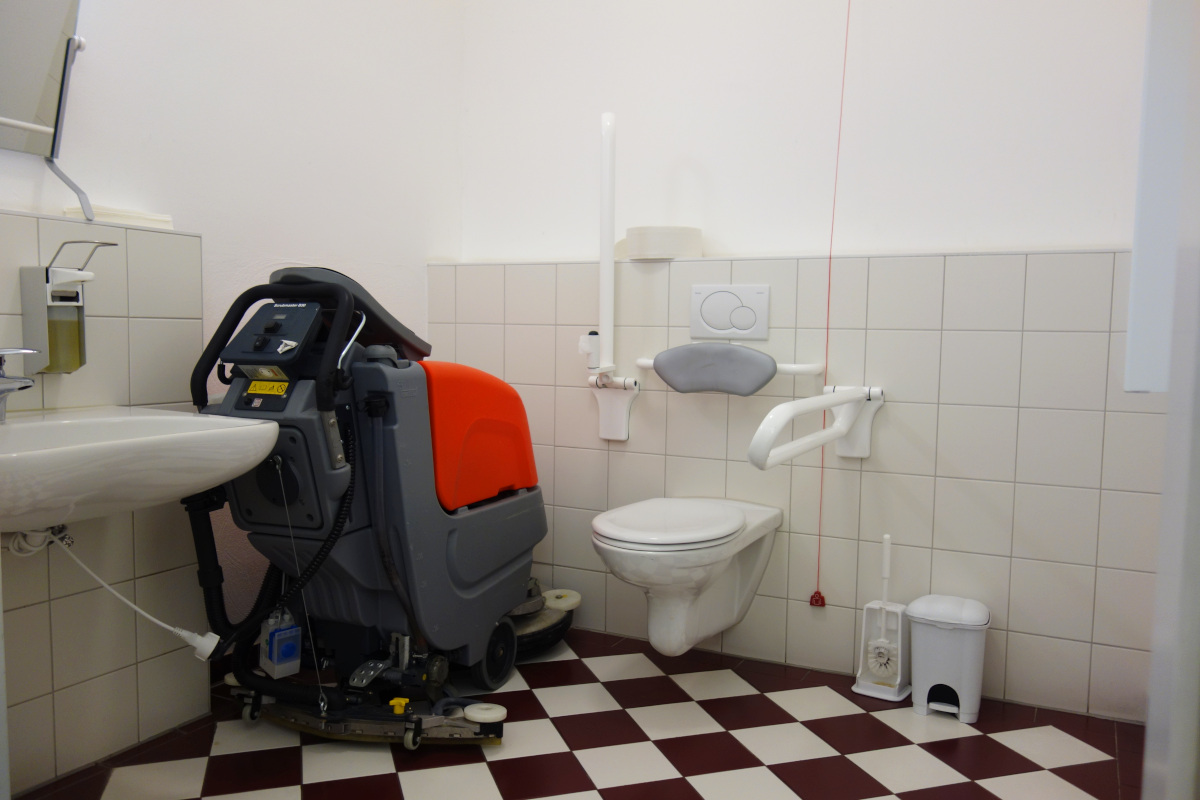 Das Behinderten-WC mit unterfahrbarem Waschbecken und Stützgriffen neben dem WC sowie einer Reinigungsmaschine im Eck des WCs.