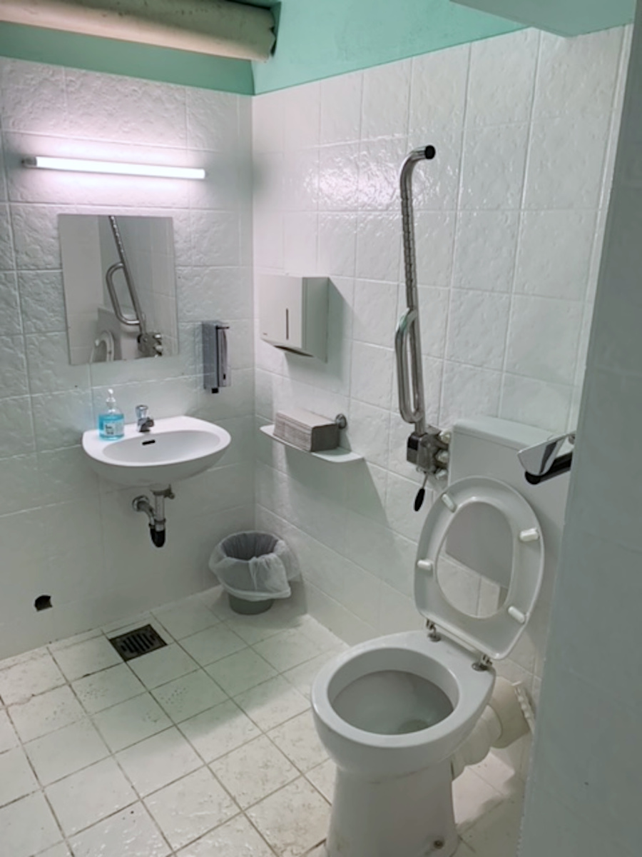 Behinderten-Toilette. Alles weiß gefliest. Toilette mit einem Stütz-Griff links. Kleines Wasch-Becken im Hintergrund.