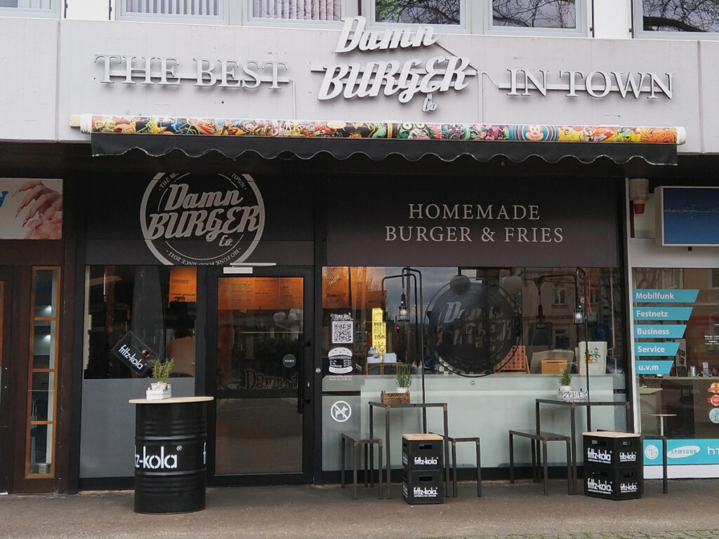 Blick von außen auf den Burgerladen mit gläserner Eingangstür und Glasfront. Über der Front befindet sich der Schriftzug „Damn Burger“.