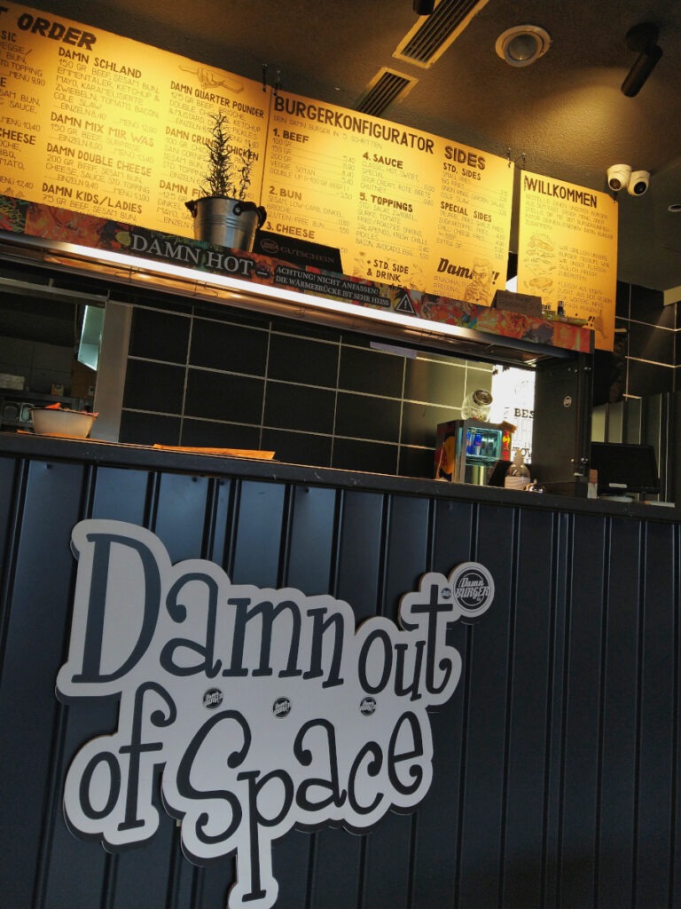 Theke mit dem Schriftzug „Damn out of Space“. Darüber ein beleuchtetes Schild mit dem Speisenangebot.
