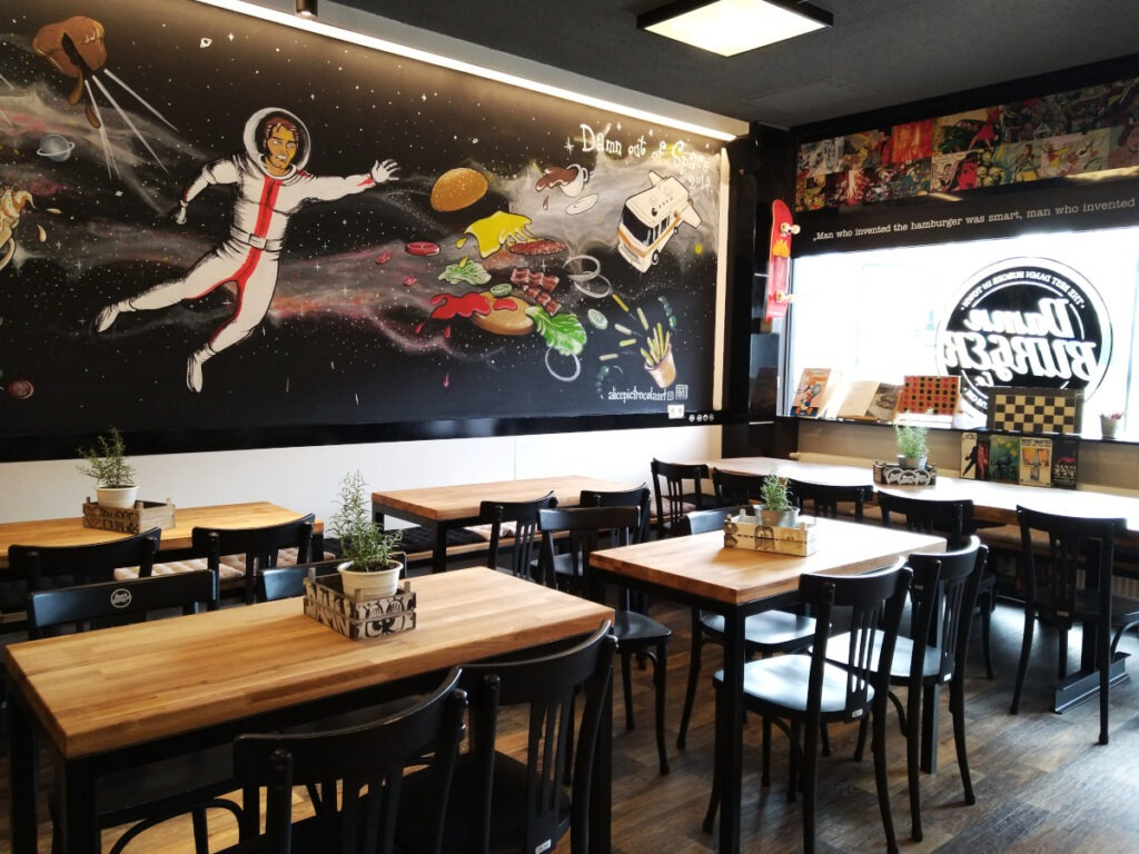 Innenbereich des Restaurants mit mehreren Vierertischen und Stühlen. Im Hintergrund eine Wandgestaltung mit einer Weltraumszene und einem Astronauten.