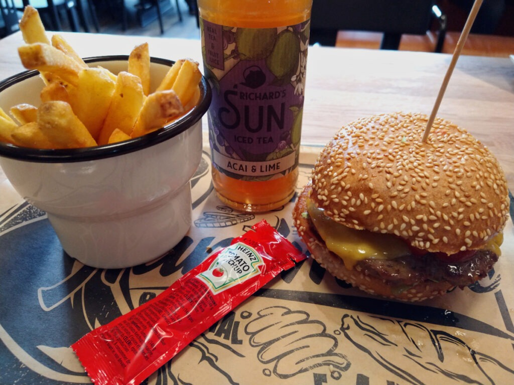Tablett mit einer Schale Pommes, einem Cheeseburger, einem Päckchen Ketchup und einer Flasche Eistee. Der Käse läuft leicht aus dem Burger.