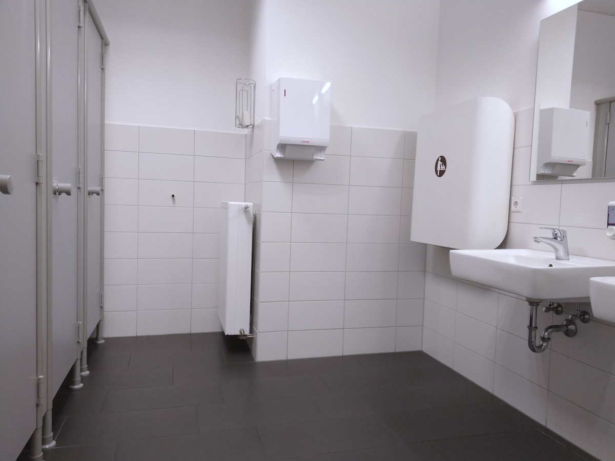 Toilette mit hellen Wänden und dunklem Boden. Links 3 WC-Kabinen. Rechts ein Wickel-Tisch und 2 Wasch-Becken.