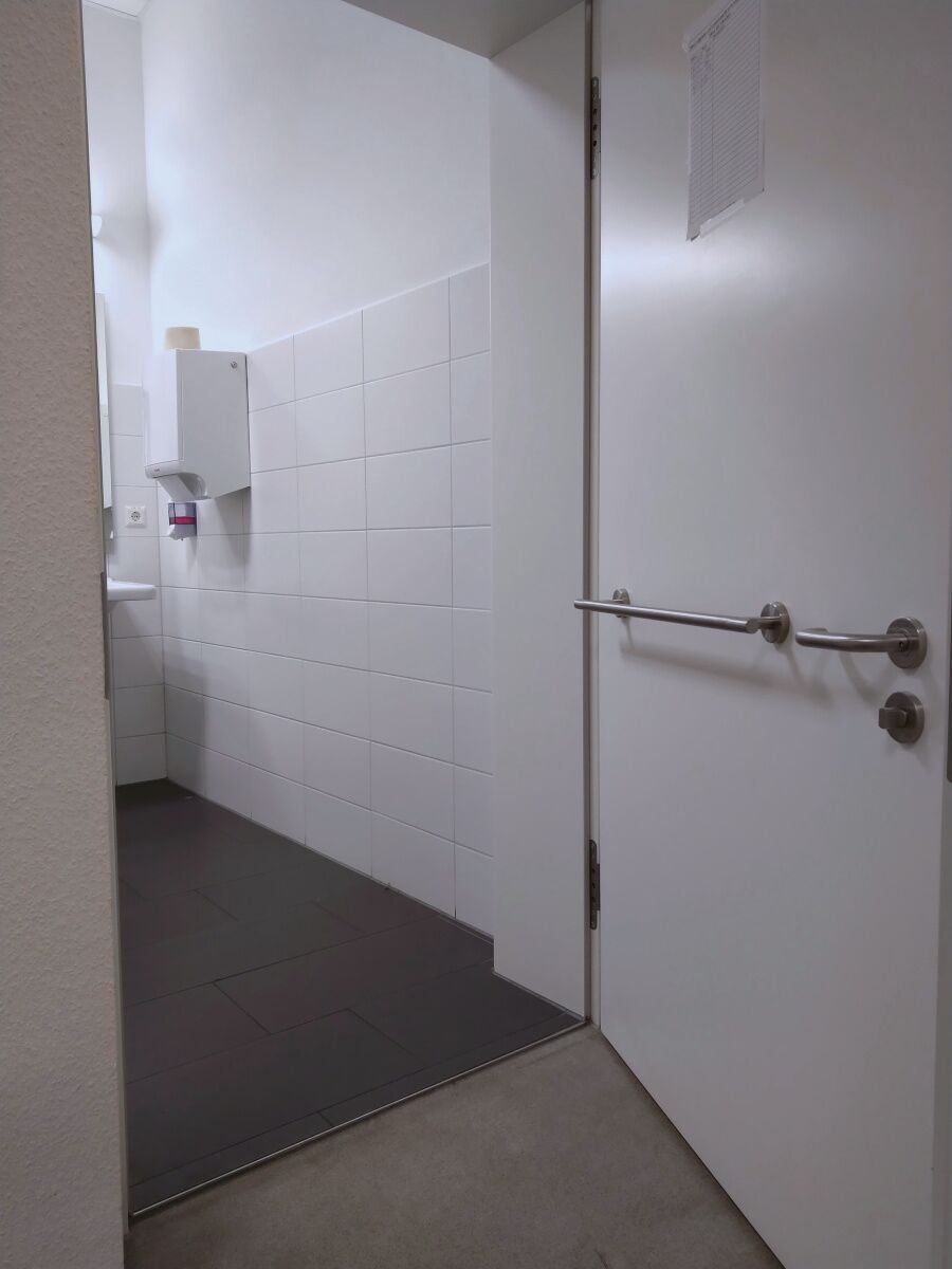 Geöffnete Tür zur Behinderten-Toilette. Auf der Innen-Seite der Tür ein Zuzieh-Stangen-Griff.