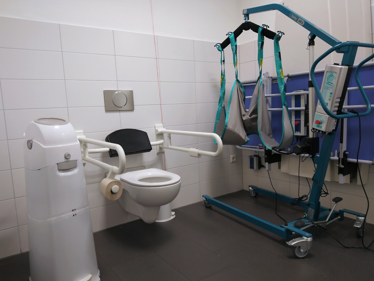 Die Behinderten-Toilette hat 2 klappbare Stütz-Griffe. Links vom WC ein Mülleimer. Rechts ein Personen-Lifter und eine eingeklappte Liege.