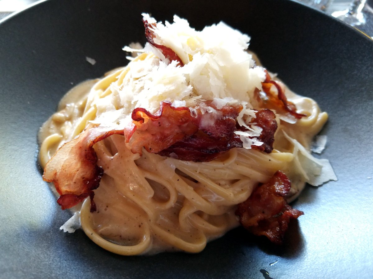 Linguine Carbonara in Nahaufnahme. Pasta glänzend durch das Eigelb. Auf den Nudeln kross gebratener Speck und gehobelter Käse.