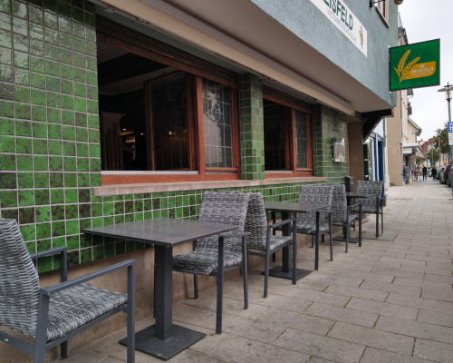 Außen-Ansicht vom Restaurant "Im Reisfeld". Außen-Wand mit grünen Fliesen und 2 Fenstern mit Bunt-Glas. Davor 3 Tische mit jeweils 2 Stühlen.