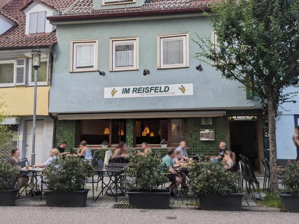 Mehrere Tische und Stühle mit Gästen im Außenbereich. Die Tische zur Straße sind durch bepflanzte Kübel abgetrennt. Im Hintergrund ist das Restaurant mit einem großen Wandschild mit der Aufschrift „Im Reisfeld“ zu sehen.
