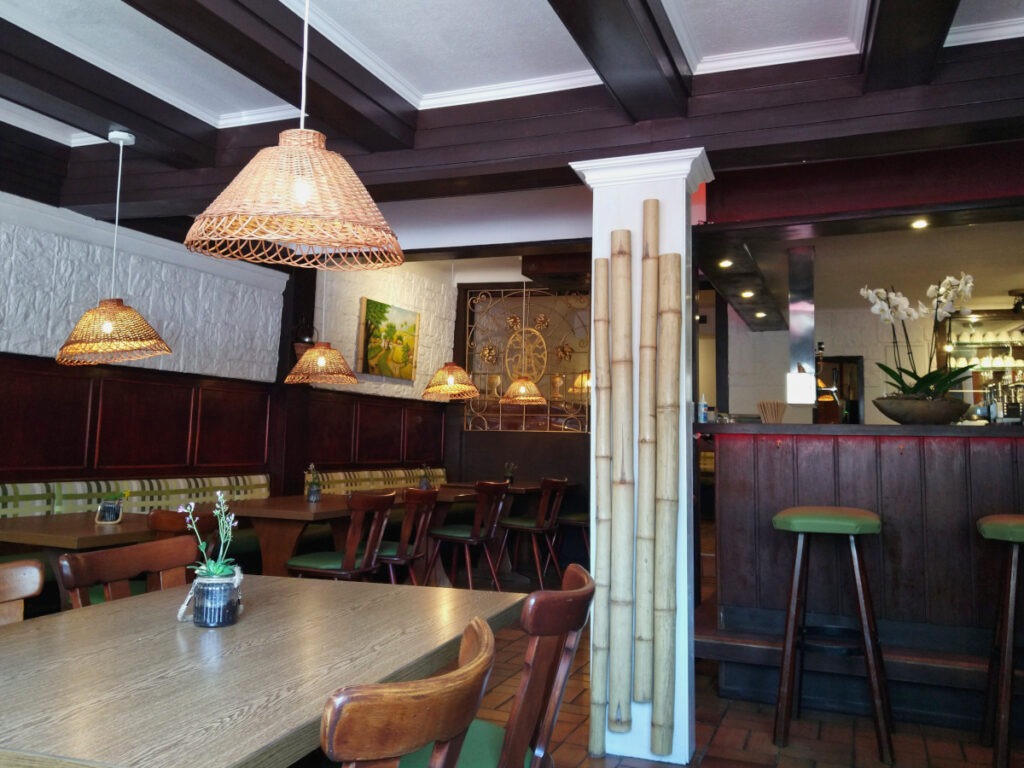 Innenbereich des Restaurants mit mehreren Tischen entlang der Wände. An den Wänden befinden sich Sitzbänke, gegenüber stehen Stühle. Rechts ist eine Theke, schräg daneben eine Säule.