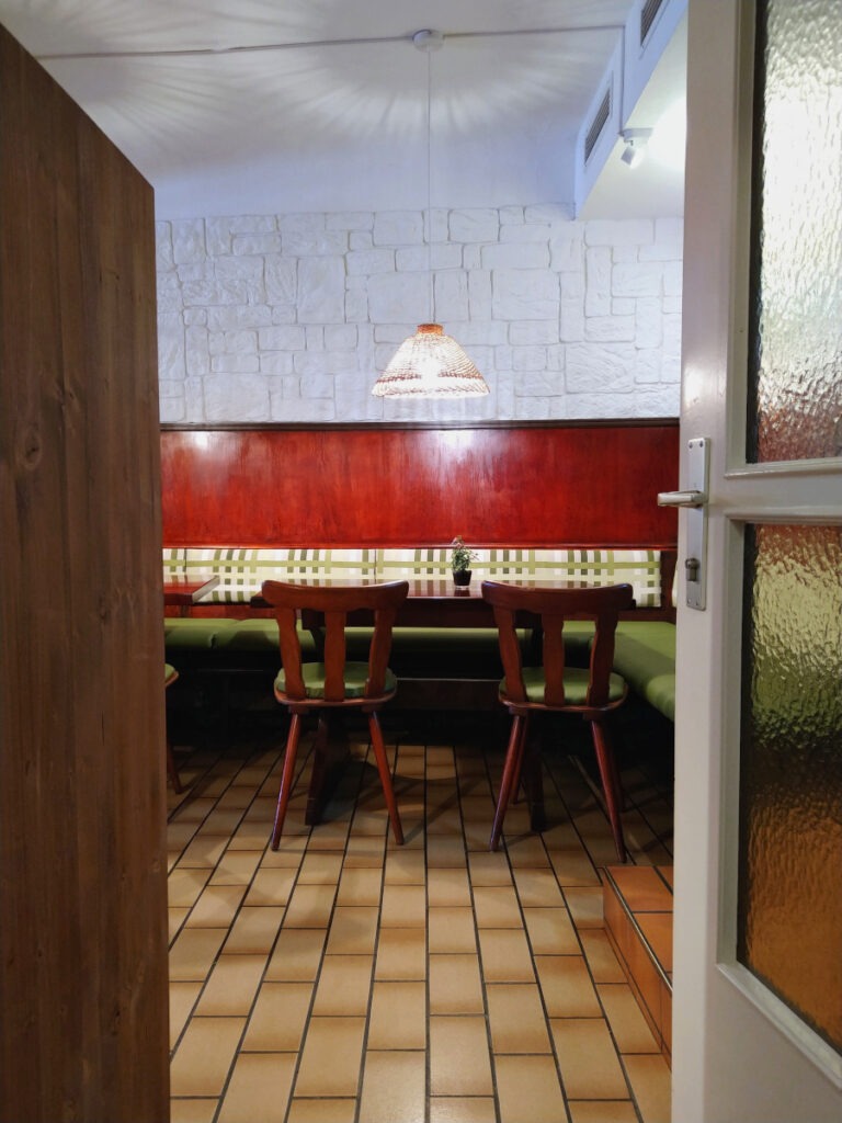 Blick durch eine geöffnete Tür in den hinteren Restaurantbereich mit Sitzbänken entlang der Wand. Im Vordergrund steht ein Tisch mit zwei Stühlen.