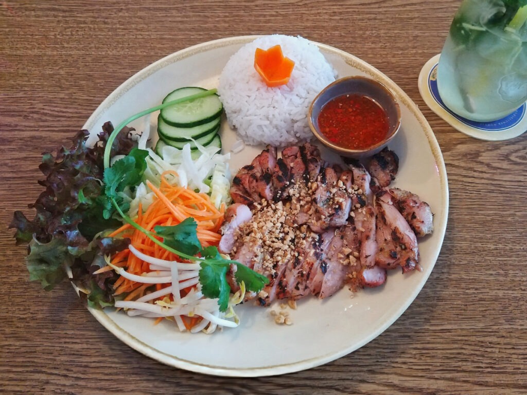 Teller mit in Streifen geschnittenem Fleisch. Daneben eine halbkugelförmige Portion Reis sowie gestiftelte Karotten und Kohlrabi. Dazu eine Schale mit roter Würzsoße.