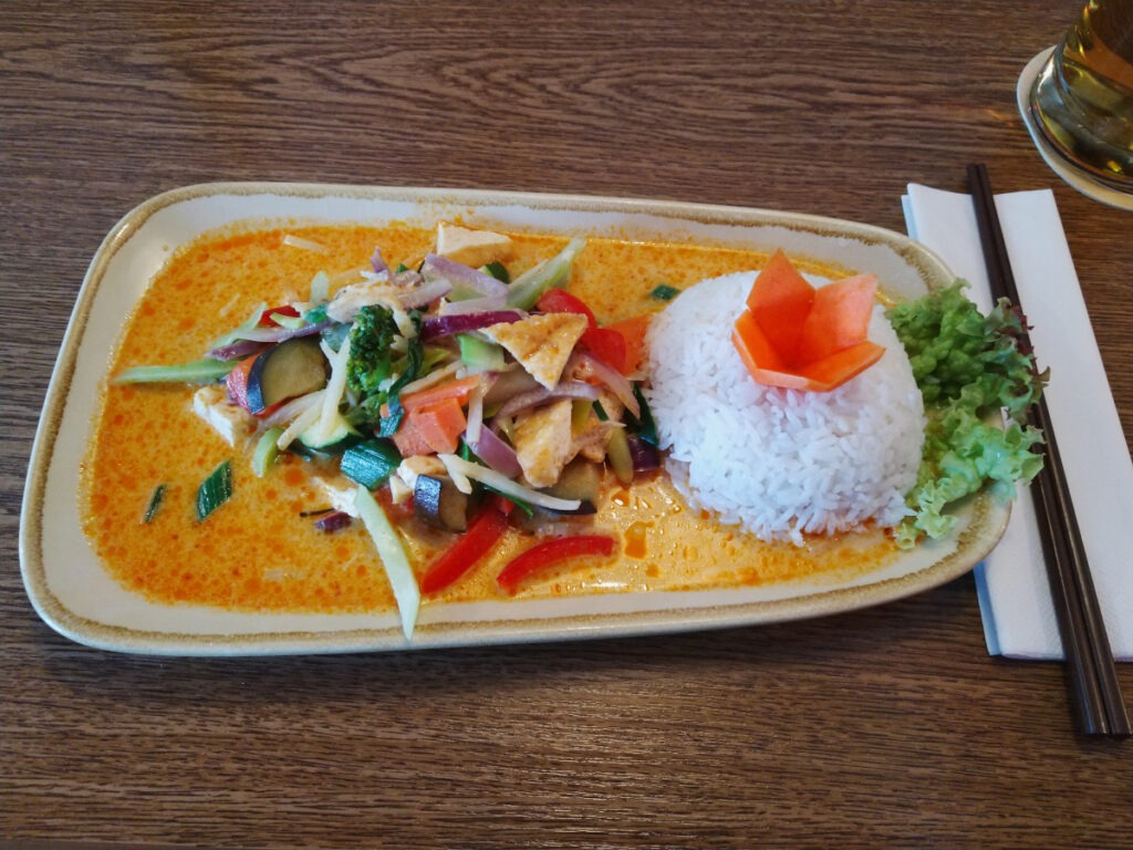 Länglicher Teller mit gelblich-roter Soße. Rechts eine halbkugelförmige Portion Reis, links daneben Tofustücke und verschiedenes, in Stifte geschnittenes Gemüse.