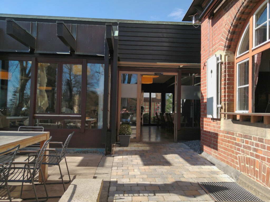 Eingang des Restaurants mit elektrisch öffnenden Glastüren, links ein Teil der Außenterrasse mit einem Tisch und Stühlen.