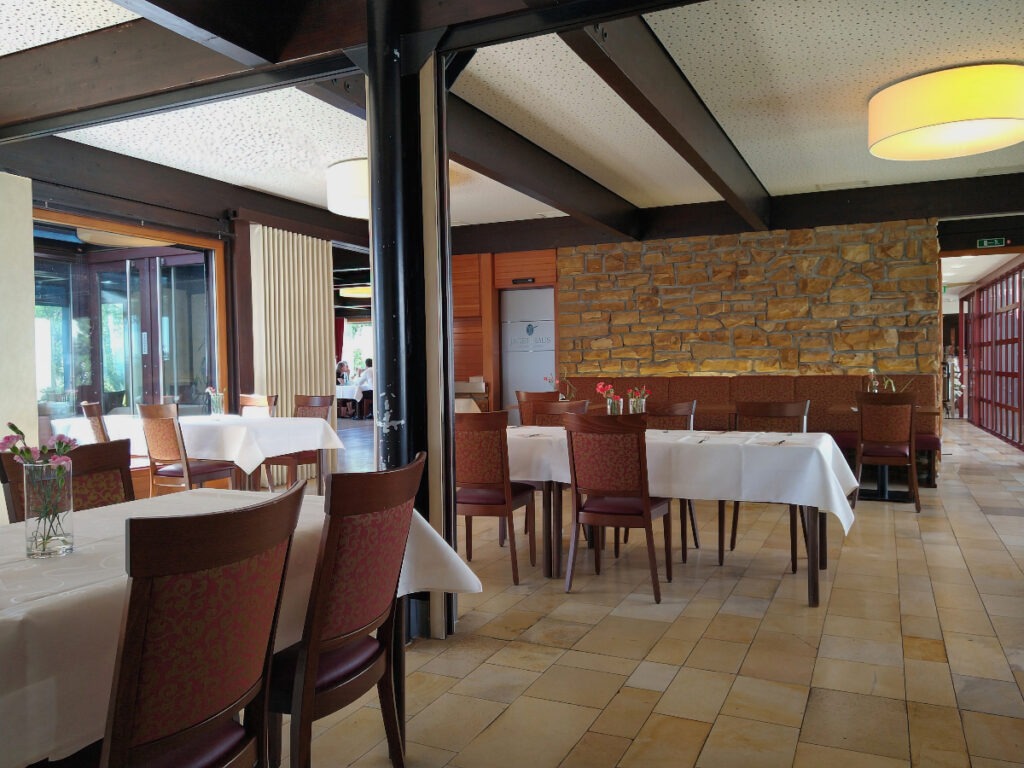 Innenbereich mit glattem Steinboden, mehreren Tischen mit Tischdecken und Stühlen. Große Fensterfronten und eine Seitenwand in Natursteinoptik.