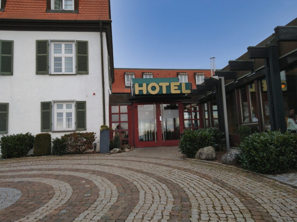 Gepflasterte Fläche vor dem Eingang mit zweiflügeliger Glastür und breitem roten Rahmen.