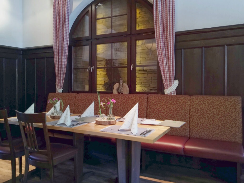 Zwei Tische mit Stühlen und Sitzbank an der Wand. Auf dem Tisch Platzdecken, Besteck und aufgestellte Servietten. Wände mit dunkler Holzverkleidung.