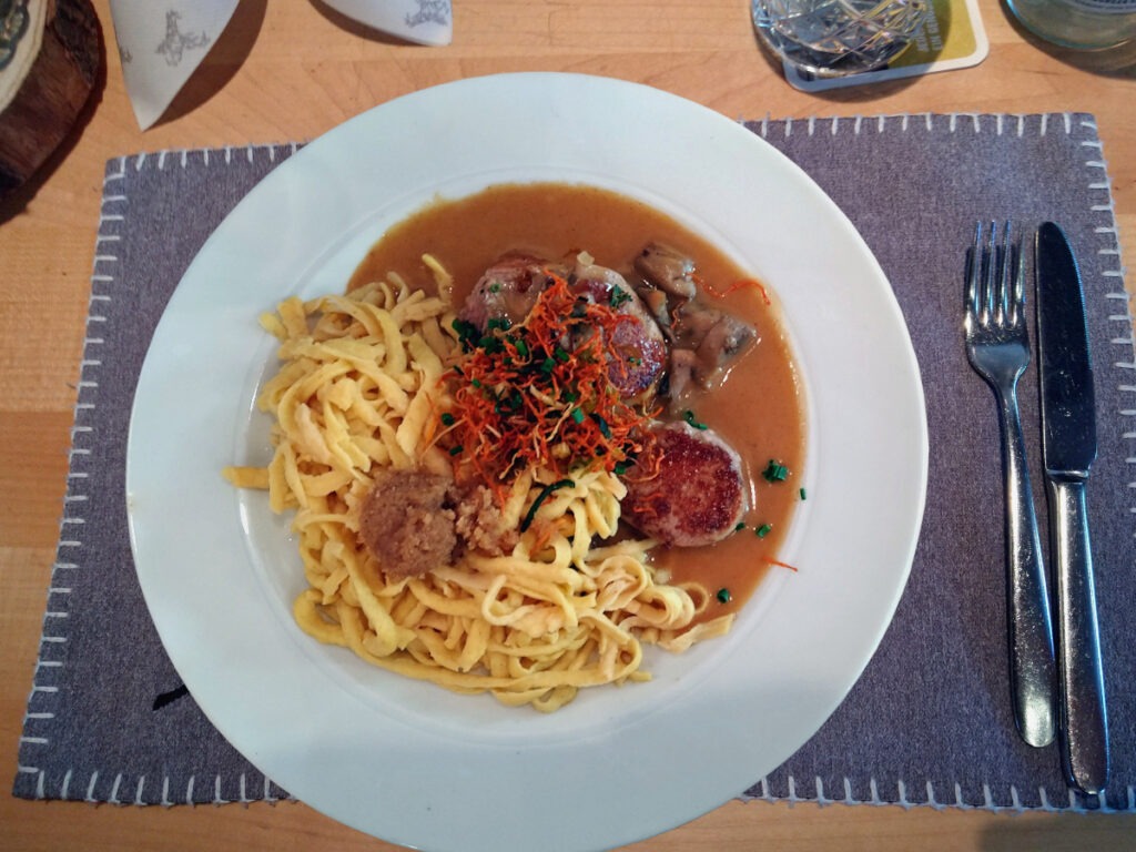 Teller mit großer Portion Spätzle, daneben mehrere Scheiben Schweinelende mit brauner Soße, dazu einige Pilze.