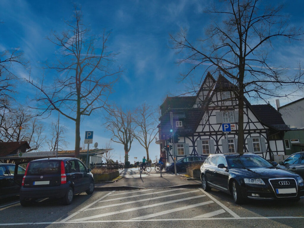 Parkplatz mit links und rechts geparkten Autos. In der Mitte führt ein Weg zu einer Fußgängerampel, auf der gegenüberliegenden Straßenseite befindet sich das Restaurant Jägerhaus.