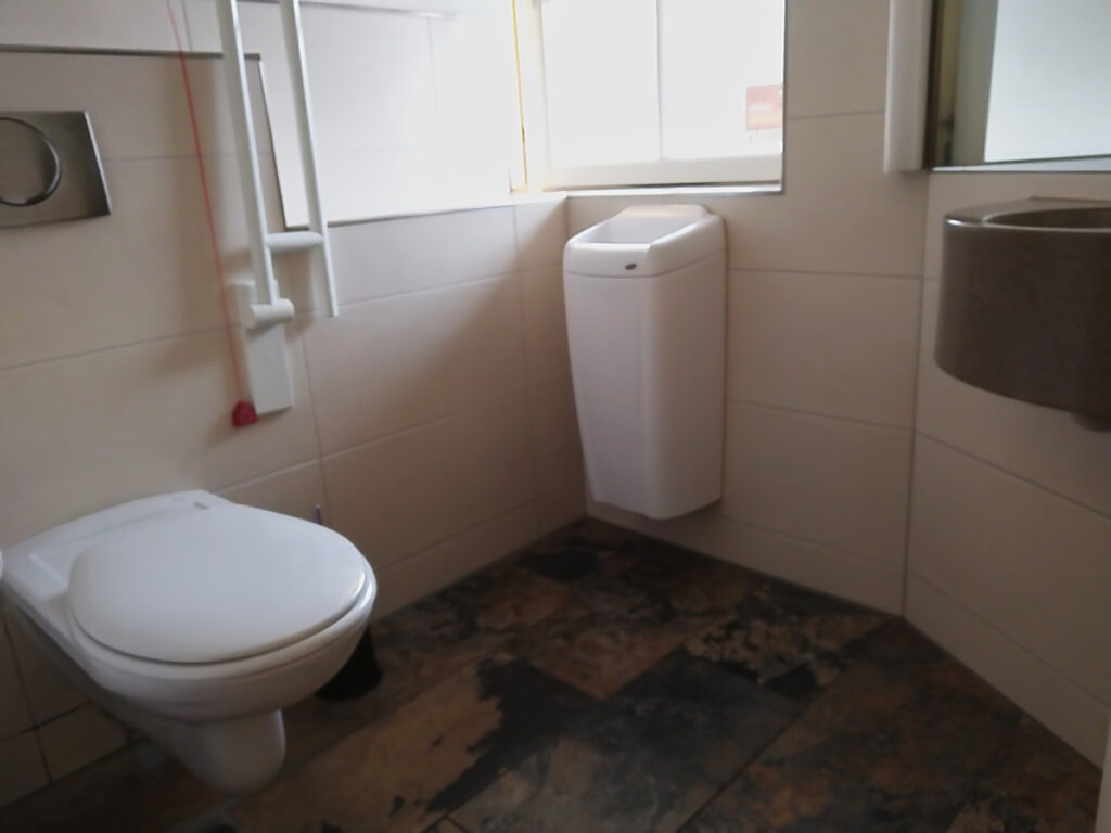 Beengte Platzverhältnisse in der Toilette mit WC und klappbarem Haltegriff links. Rechts daneben ein Mülleimer und an der Wand ein kleines Waschbecken.