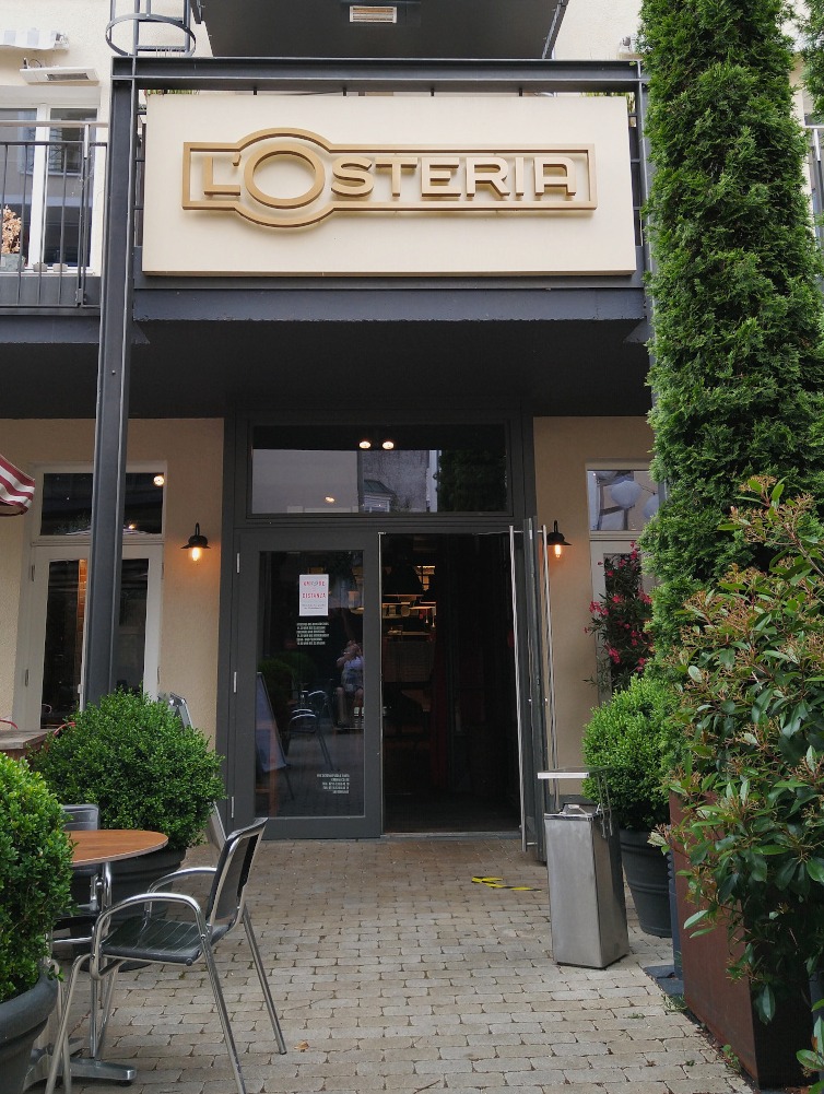 Geöffnete Glastür mit Metallrahmen und Blick in das Restaurant. Links stehen ein Tisch mit einem Stuhl, daneben mehrere Blumenkübel mit buschigen Pflanzen.