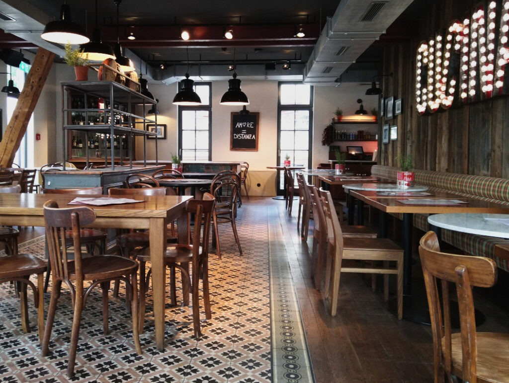 Innenbereich eines Restaurants mit mehreren Tischen und Stühlen links. Rechts Tische mit Sitzbank entlang der Wand und gegenüberliegend Stühlen. Gemütliche und stilvolle Inneneinrichtung.