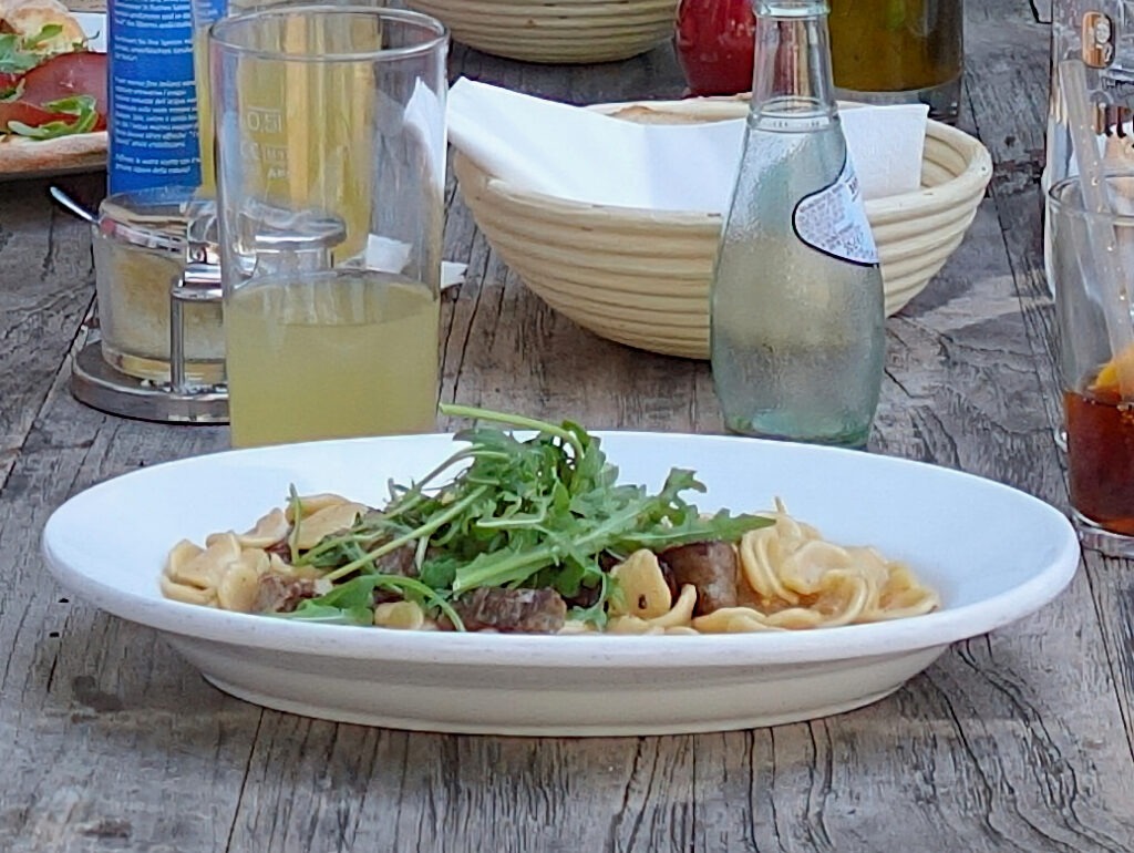 Teller auf rustikalem Holztisch mit Pasta, Fleischstücken und Rucolablättern.