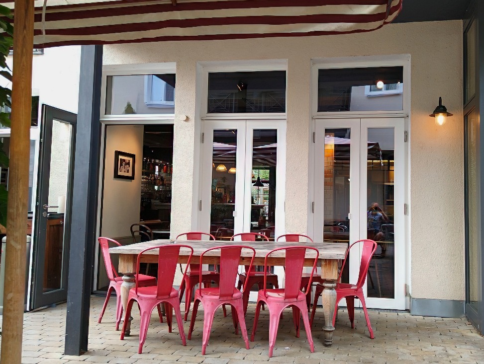 Holztisch mit roten Metall- oder Plastikstühlen auf einer Terrasse, im Hintergrund eine geöffnete Tür zum Innenbereich des Restaurants.