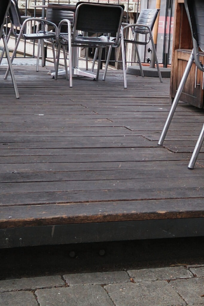 Großaufnahme einer Stufe im Außenbereich, im Hintergrund auf einem erhöhten Terrassenbereich stehende Tische und Stühle.