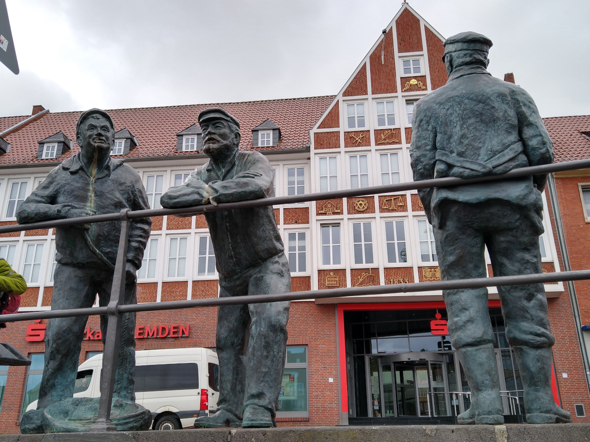 3 lebens-große Bronze-Figuren von Männern, die lässig an ein Geländer lehnen.