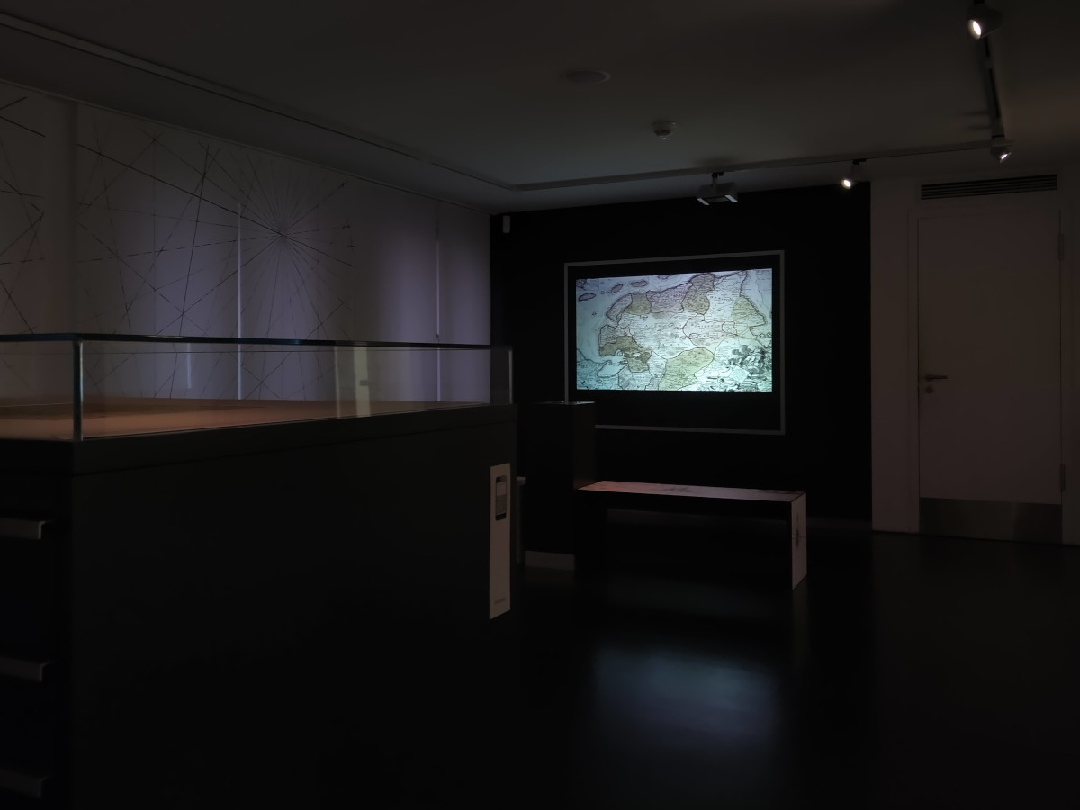 Dunkler Raum mit einem großen Schubladenschrank und einem Fernseher, auf dem eine historische Karte von Ostfriesland zu sehen ist.