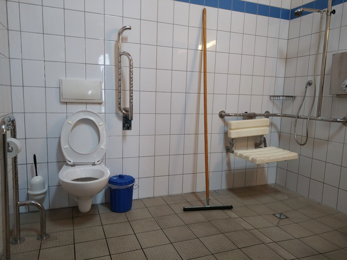 Gefliester Sanitär-Raum. Auf der linken Seite eine Behinderten-Toilette mit Klapp-Griff. Auf der rechten Seite eine Dusche mit Dusch-Sitz.