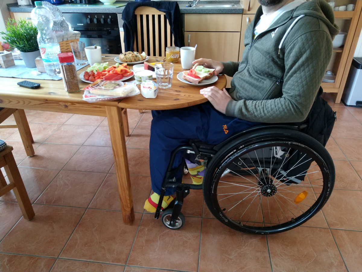 Fürs Frühstück gedeckter Tisch. Am Tisch ein Rollstuhlfahrer beim Frühstücken.