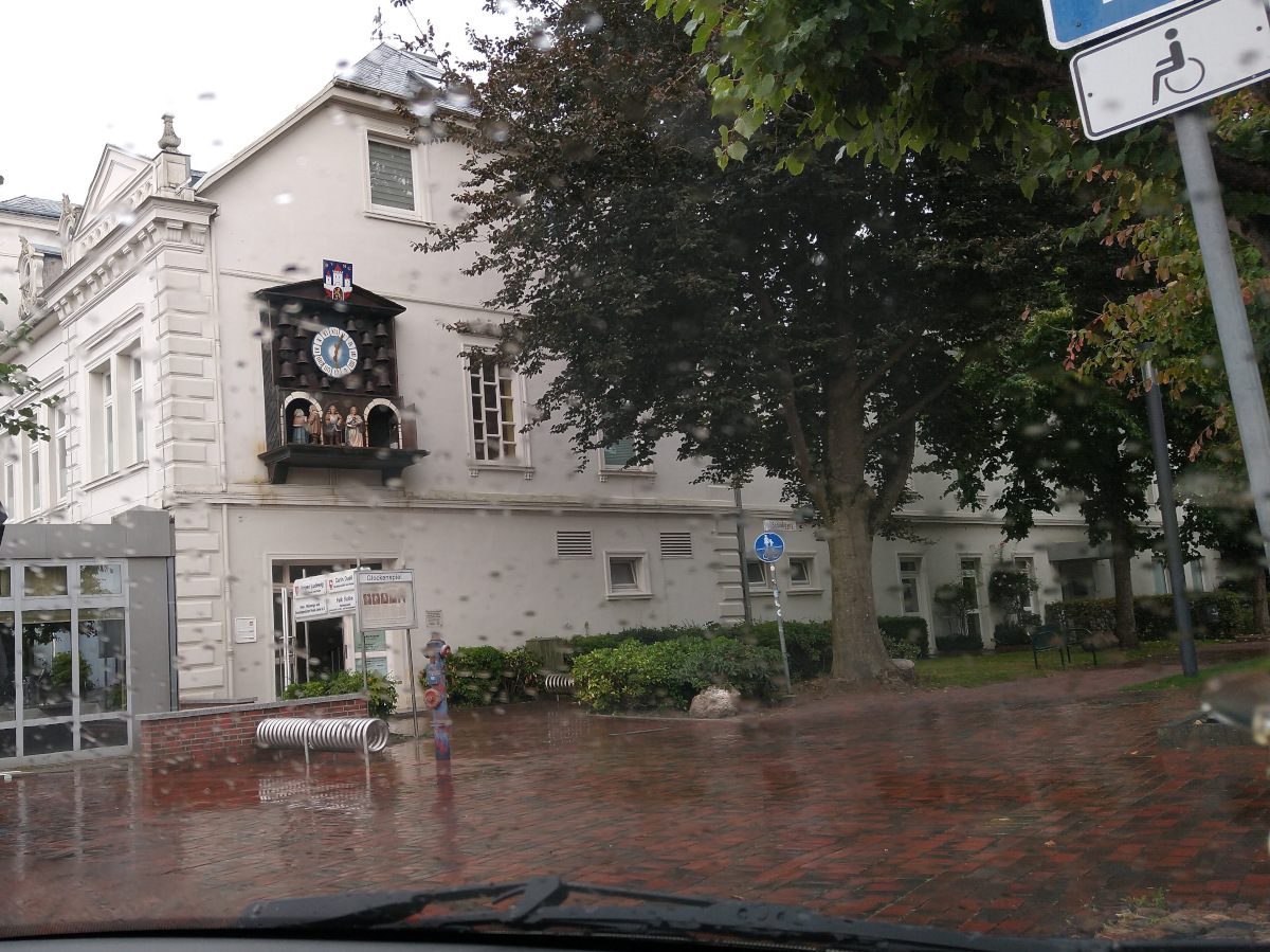 Blick aus einem Auto. Draußen Regen. Links am Gebäude das Glockenspiel mit Figuren.
