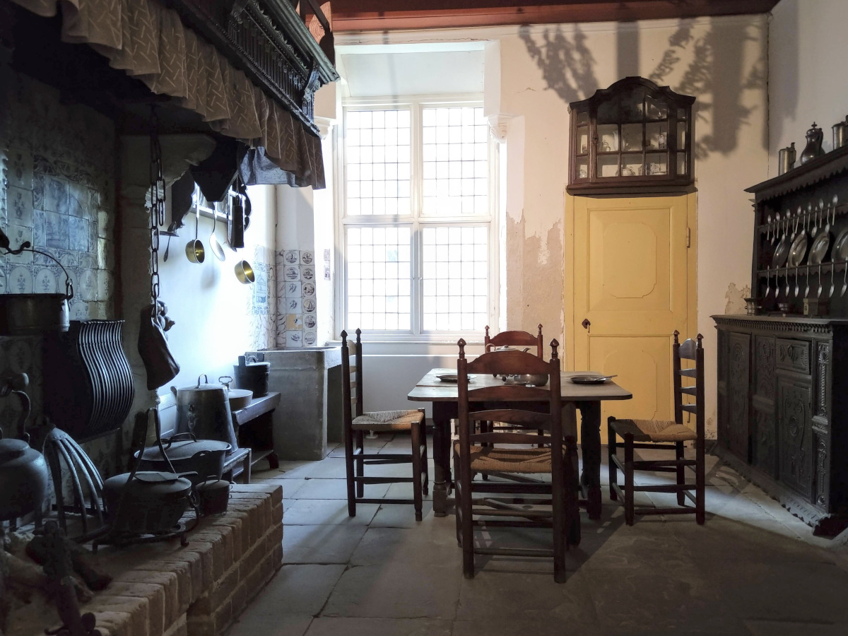 Historische Küche: Links eine offene Feuerstelle mit Töpfen. Mittig ein Holz-Tisch mit Stühlen. Rechts ein verzierter Schrank mit Geschirr.
