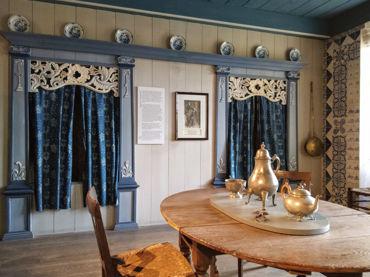Im Vordergrund Tisch mit historischem Tee-Service aus Blech. Im Hintergrund Schlaf-Kammern, die mit einem Vorhang abgetrennt sind.