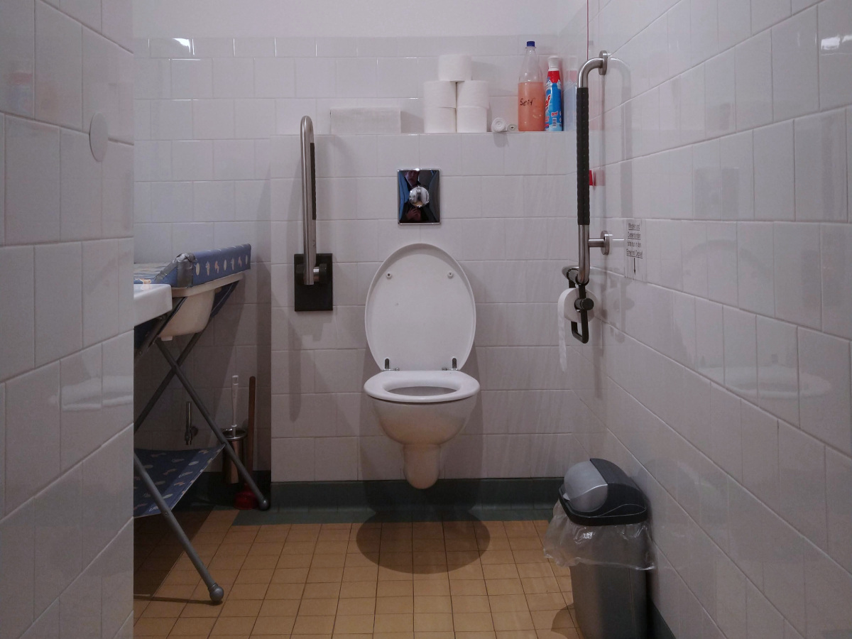 Kleine Behinderten-Toilette. Links eine Wickel-Liege und ein Waschbecken. Mittig eine Toilette mit einem Klapp-Griff und einem Griff an der Wand.