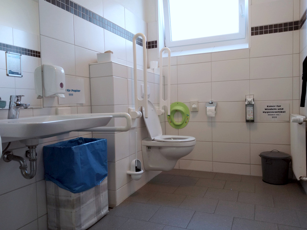 Behinderten-Toilette: länglicher Raum, links vorne das Waschbecken, links hinten die Toilette mit Stütz-Griffen.