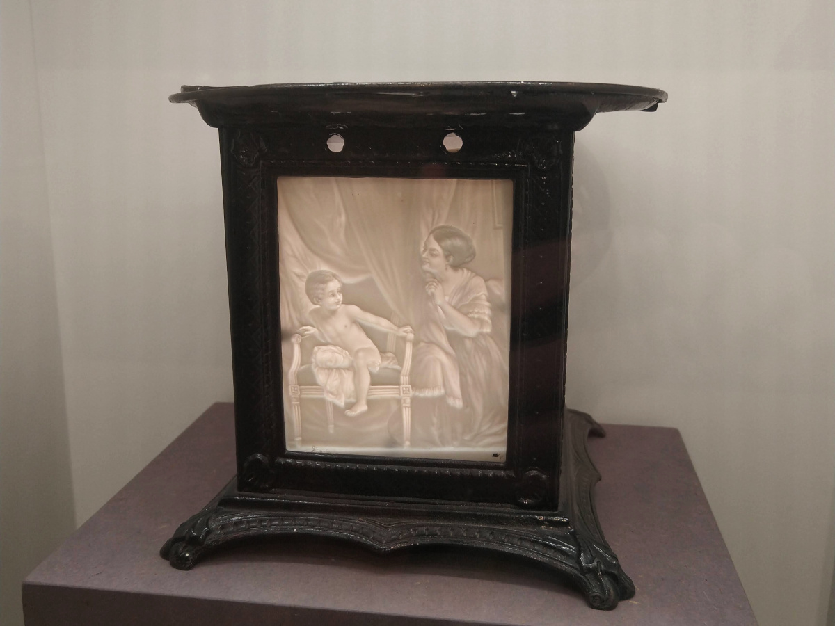 Kunstvolles Stövchen: Metall-Rahmen mit einer durchschimmernden Porzellan-Platte, auf der ein Kleinkind und eine Frau dargestellt sind.