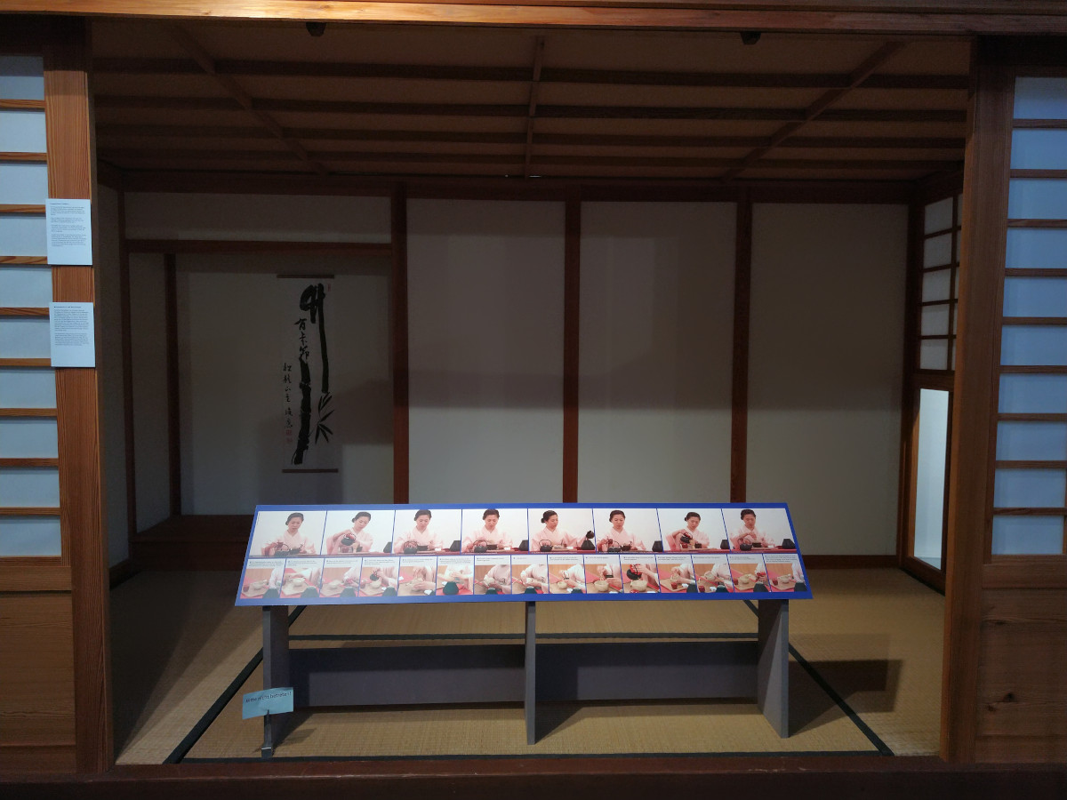 Japanische Holz-Hütte mit geöffneter Tür. Innen Stroh-Matten. Auf dem Boden eine Info-Tafel mit den verschiedenen Schritten der japanischen Tee-Zeremonie.
