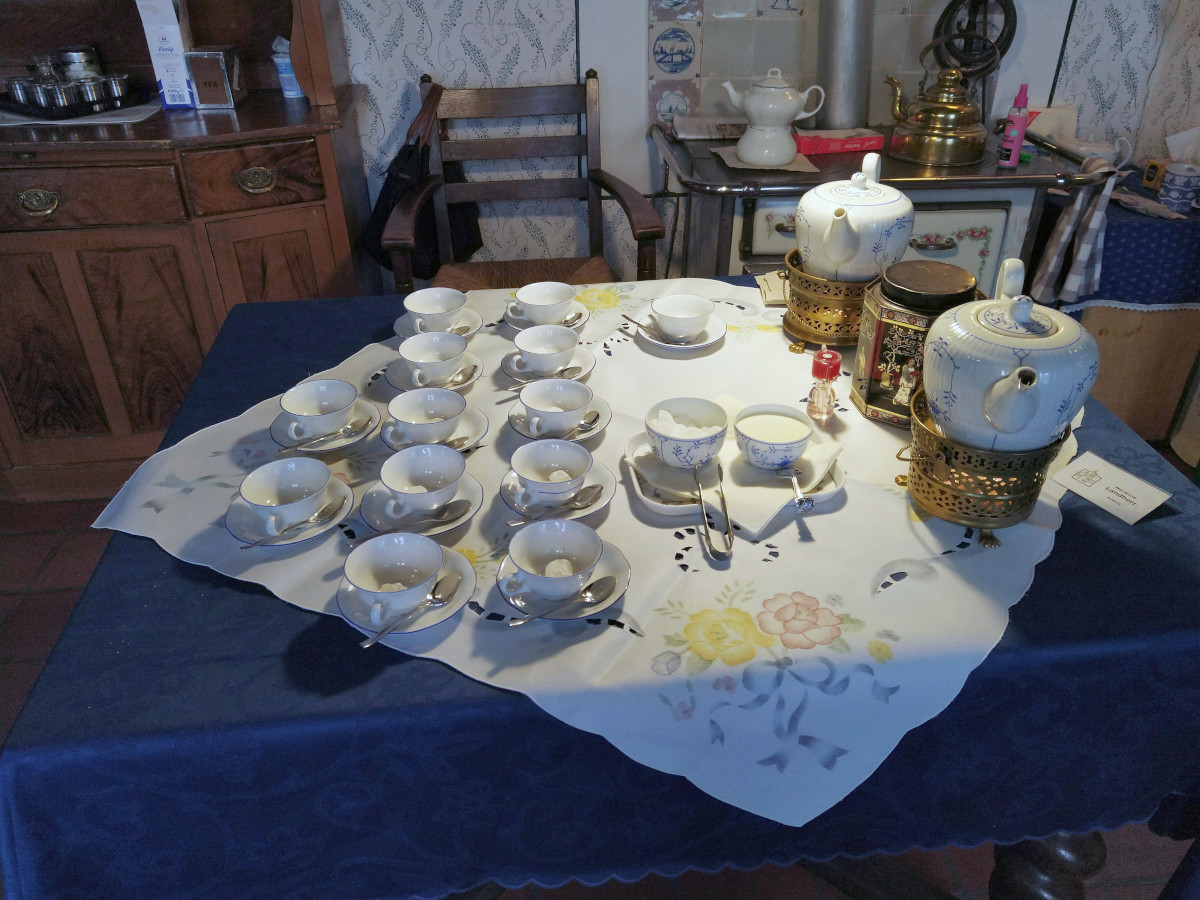 Tisch mit 13 Tee-Tassen auf Unter-Tassen. Daneben 2 große Tee-Kannen sowie ein Kandis-Schälchen und eine Tasse mit Sahne.