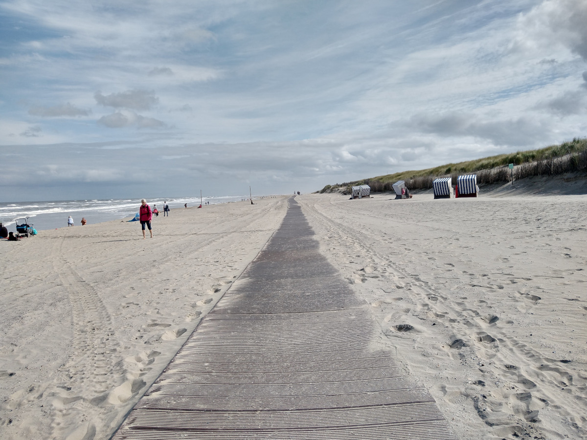 Sand-Strand. In der Mitte ein langer Kunststoff-Steg entlang vom Strand. Links am Rand Meer. Rechts einzelne Strand-Körbe.